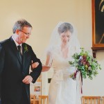 A bride holds her father's arm and is looking down. She is wearing a white dress, veil and is holding a bouquet of flowers. The father is wearing a dark suit, white shirt and tie.